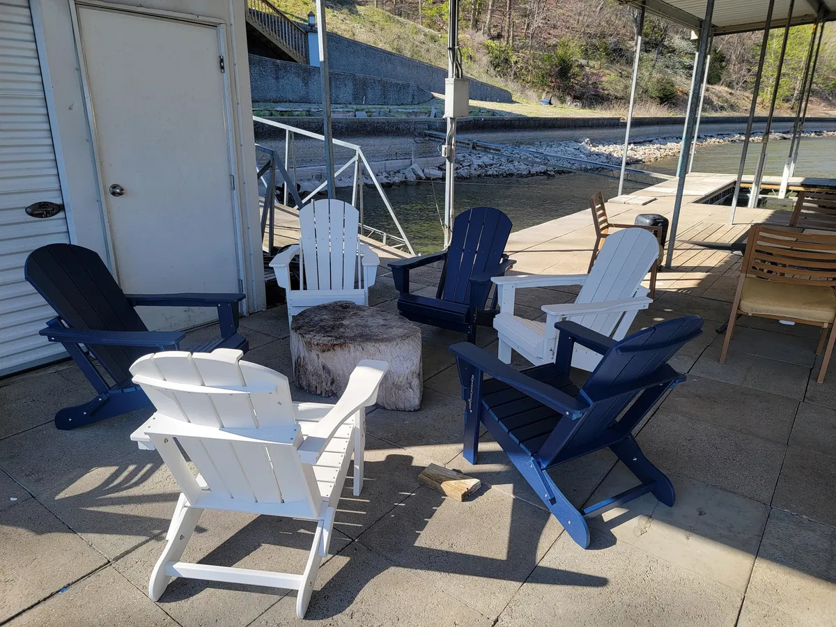 A relaxed waterfront setting with Adirondack chairs arranged around a wooden stump, facing the water. The scene suggests tranquil moments spent socializing, watching sunsets, or stargazing by the lake’s edge.