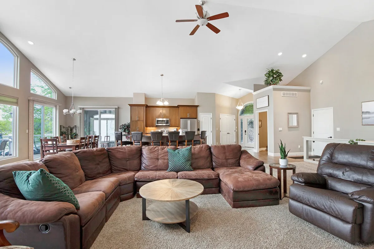 This image shows a spacious, open-concept living area featuring a large sectional sofa, a round coffee table, and a dining area that seamlessly connects to a modern kitchen. The space is bathed in natural light, creating a welcoming environment perfect for group gatherings, game nights, or relaxed conversations.