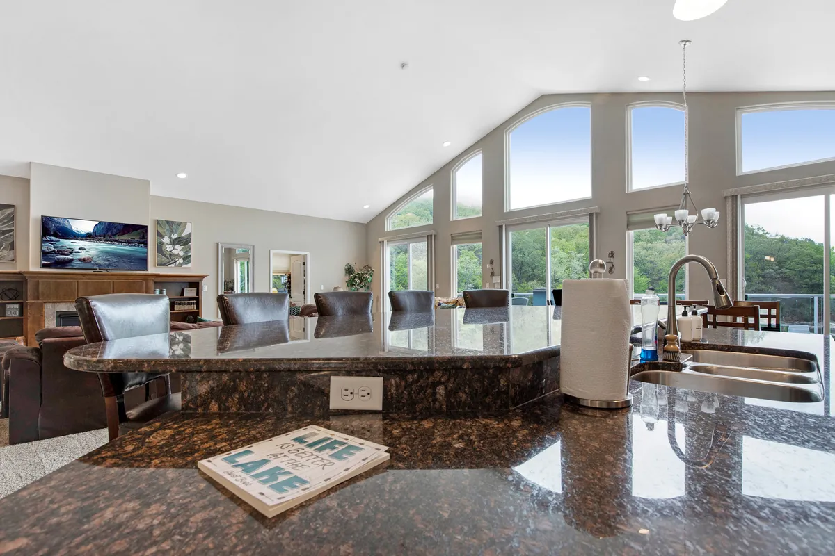 A lively interior scene featuring large windows overlooking the lake, a kitchen island ideal for group meal prep, plush seating for relaxation, and stylish artwork. The atmosphere is set for cooking, entertaining, and sharing stories—all while enjoying sunset views.