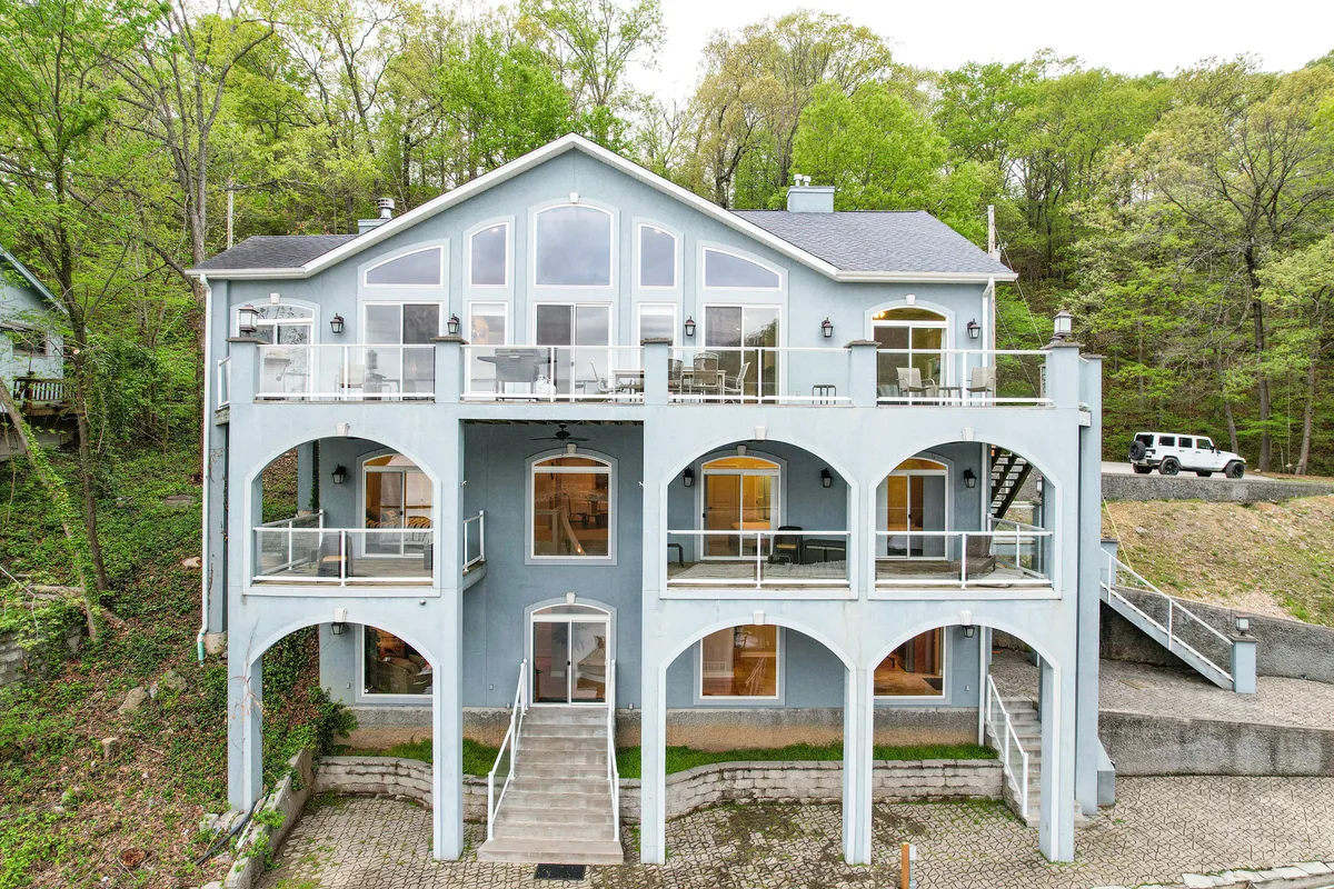 This image presents the exterior of the home, featuring large glass windows and arched balconies enveloped by lush, green trees. The architecture blends with the natural surroundings, and the generous balconies suggest ample opportunities for relaxing or socializing outdoors while appreciating the views.