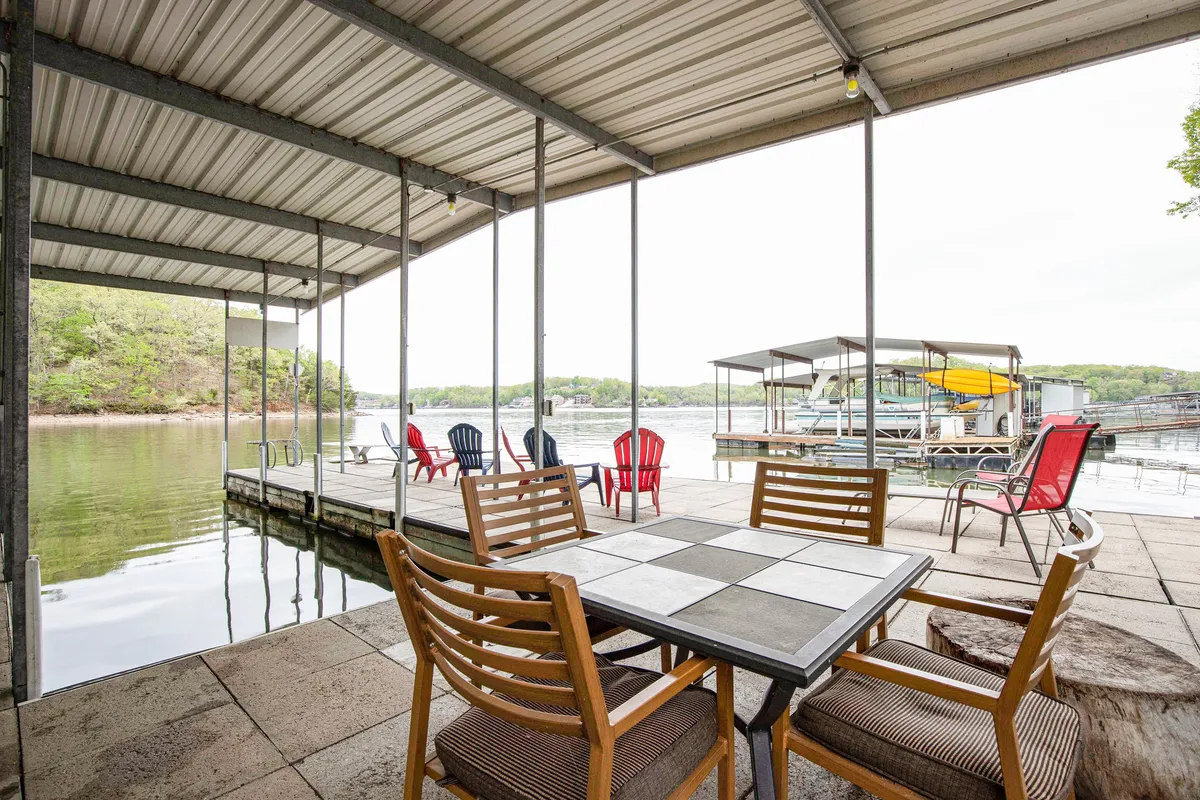 An inviting outdoor dining area with a large table and colorful Adirondack chairs sits at the lake’s edge. The waterfront view forms a stunning backdrop, suggesting leisurely meals and gatherings with the sounds of water nearby. The setup encourages both relaxed lounging and social dining.