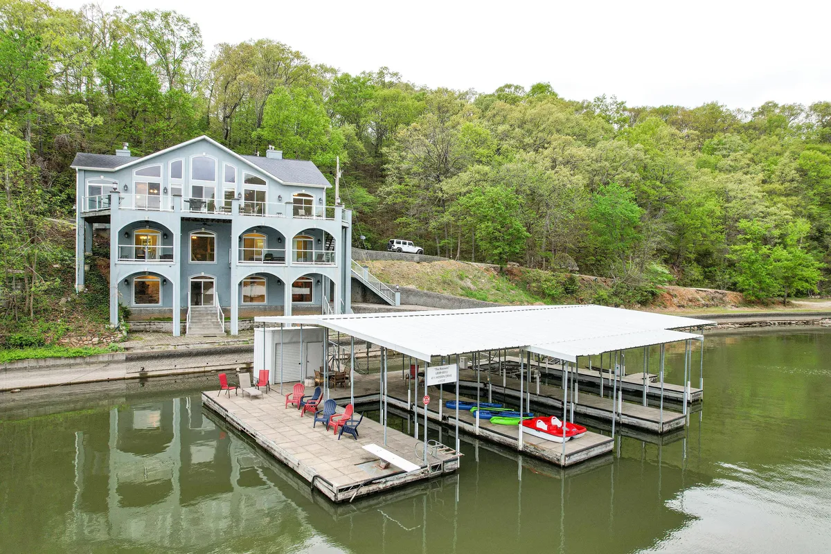 A serene lakefront scene showing the house with a dock that features boats moored on the water and lush greenery framing the property. The image captures the immediate proximity of the lake to the home, highlighting the private dock and easy access to water activities.