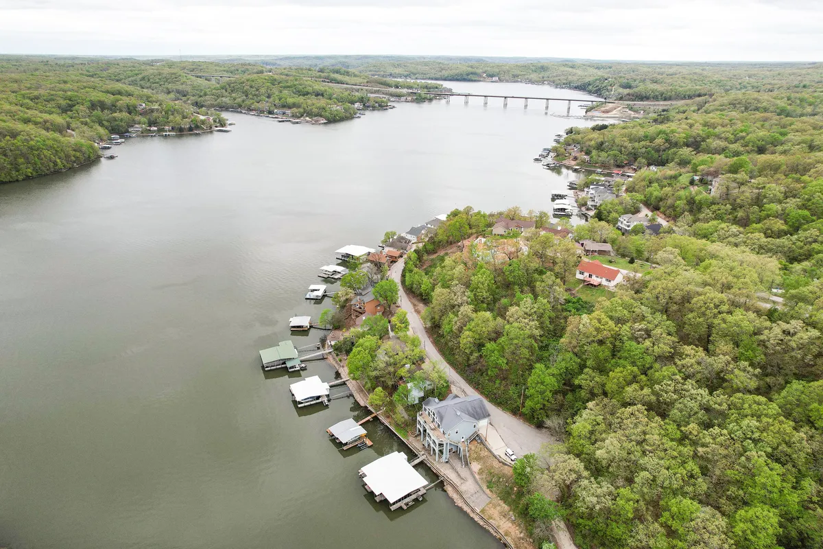 A sweeping view of the winding river lined with houses, boathouses, and bridges in the distance. The scene is tranquil, with boats potentially visible on the water and lush greenery framing the shores, evoking the community atmosphere along Lake of the Ozarks.