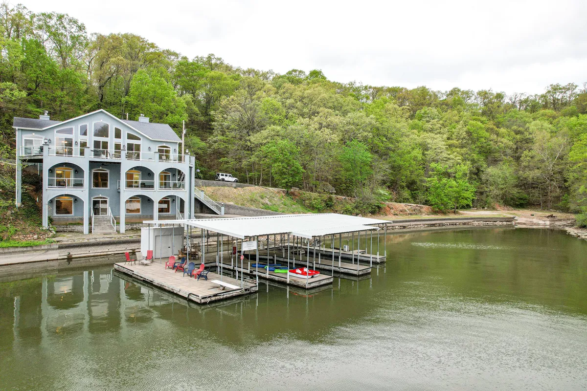 The image showcases an expansive lakeside home with floor-to-ceiling windows, nestled among trees at the edge of a tranquil cove. A private boat dock extends over calm water that reflects the lush shoreline. The scene is peaceful, suggesting both adventure and relaxation—ideal for group gatherings with easy access to water activities and sunset views.