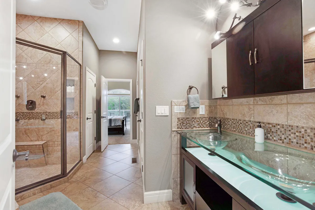 A serene, elegant bathroom featuring a glass vessel sink, walk-in shower, and decorative tilework. The design is modern and clean, suggesting luxury and meticulous upkeep.